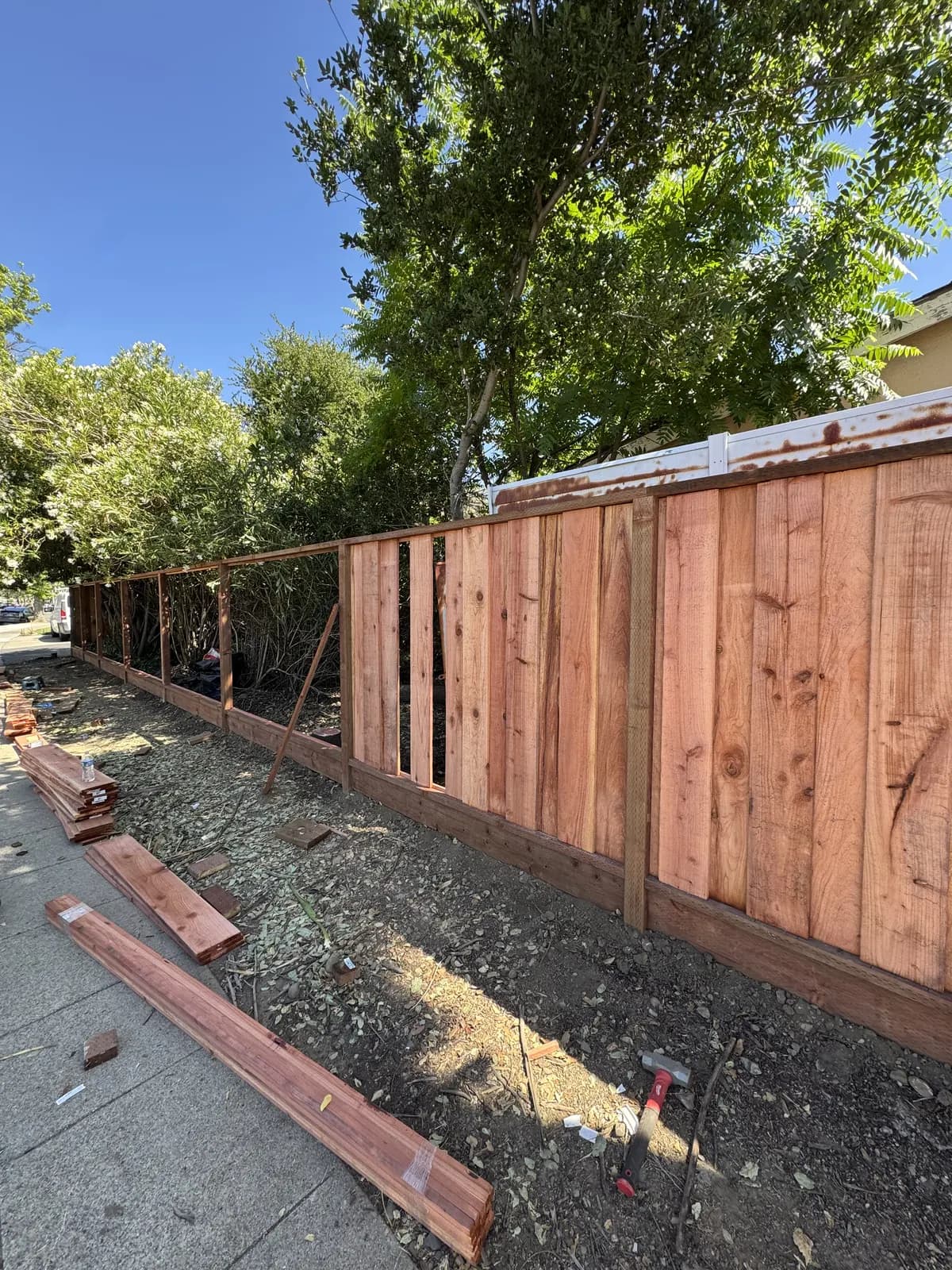 Fence run with boards fitted on the near half and bare posts and rails still open on the far half, lumber stacked on the pavement