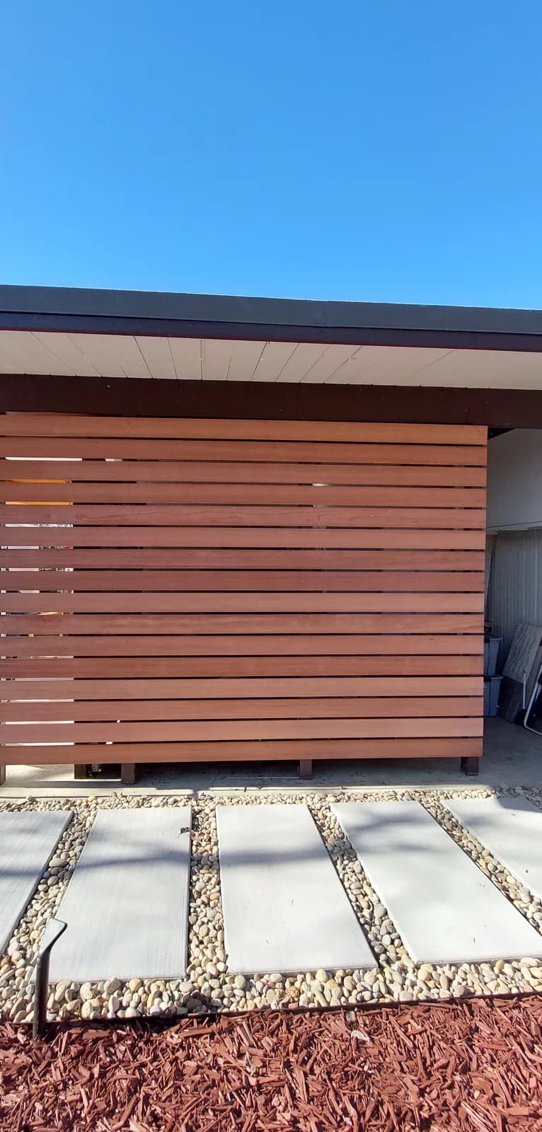 Horizontal redwood slat screen photographed square on, full height, flat roof overhang above and pebble path below
