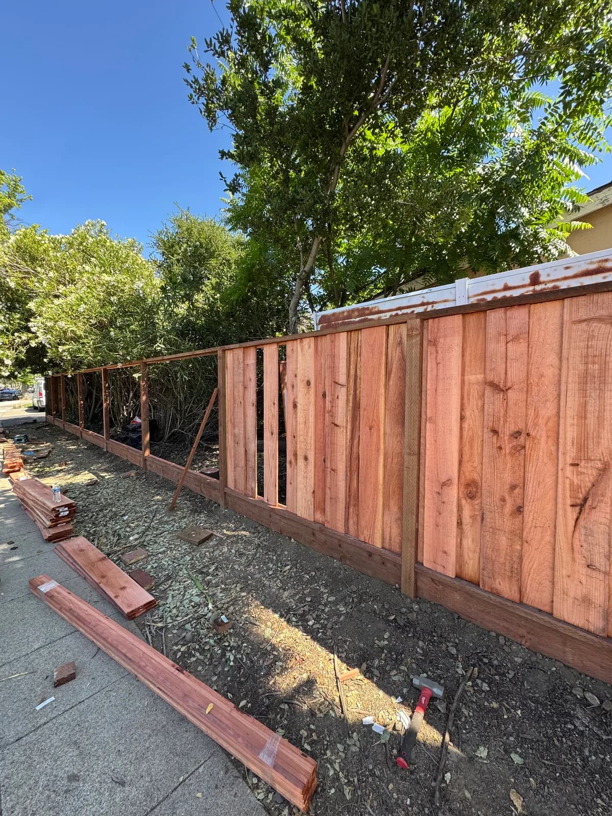 Street-side fence with boards on one half and open frame on the other, stacked redwood and a hammer on the ground