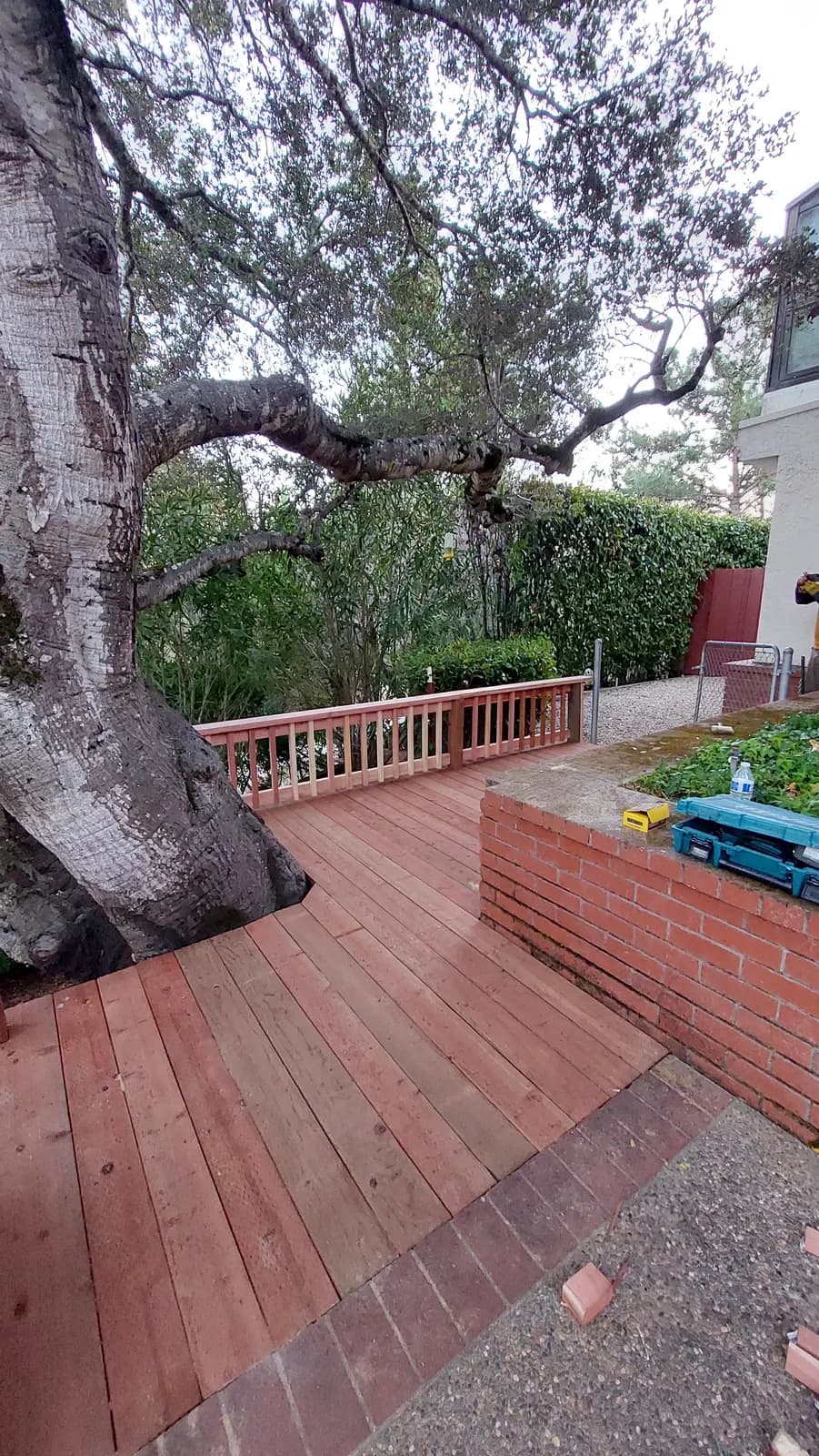 Wider view of the oak deck meeting a brick retaining wall, tool case open on the wall and a worker's drill at the edge of frame