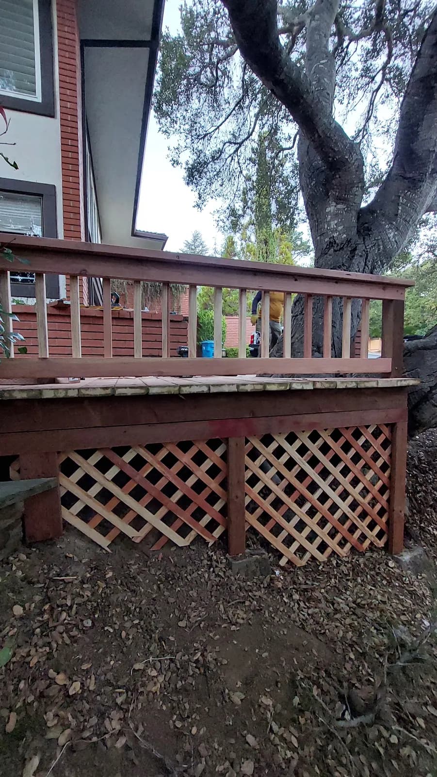 Deck edge with baluster railing above and fresh lattice skirting being fitted below, workers visible beyond