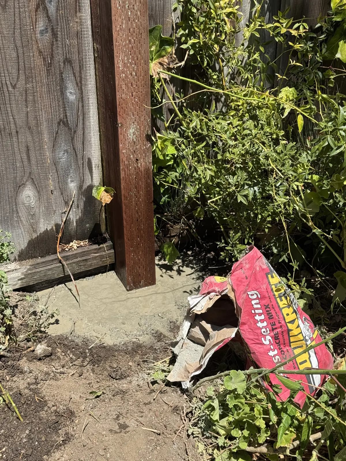 New fence post set in wet grey concrete with an opened bag of fast-setting mix lying beside the hole